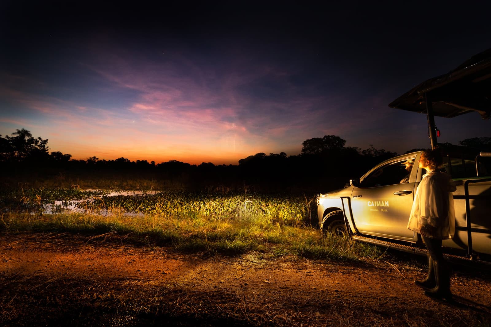 Night spotlighting starts here, at the exact minute the Pantanal stops being one thing and becomes another. A guide stands beside a Caiman lodge safari vehicle on a dirt track at dusk during a luxury Pantanal and Amazon trip, with a vivid orange and purple twilight sky reflected over a flooded marsh covered in aquatic plants