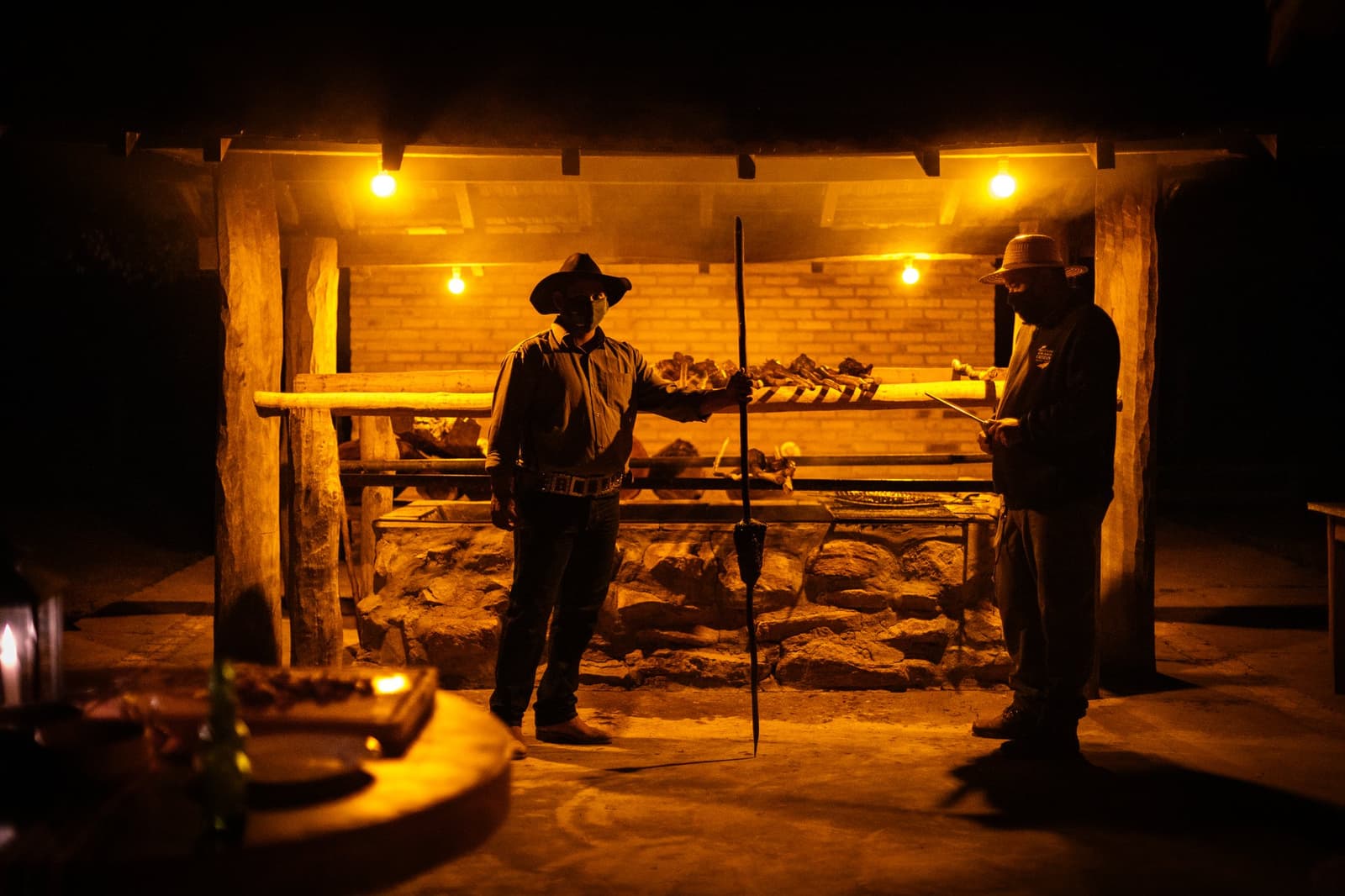Some meals earn their place at the table long before you sit down. Two gauchos in wide-brimmed hats tending large cuts of meat on a wood-fired open grill at night during a luxury Pantanal and Amazon trip, illuminated by warm amber bulbs inside a rustic stone and brick structure