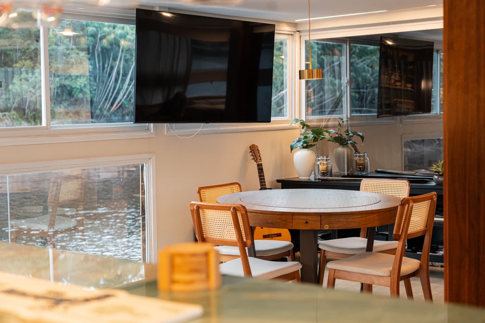 The guitar in the corner says someone on this crew knows how to end an evening. Round dining table with cane chairs inside the salon of a luxury Amazon cruise in Brazil, with a wall-mounted TV, a guitar leaning against the wall, and lush forest visible through panoramic windows