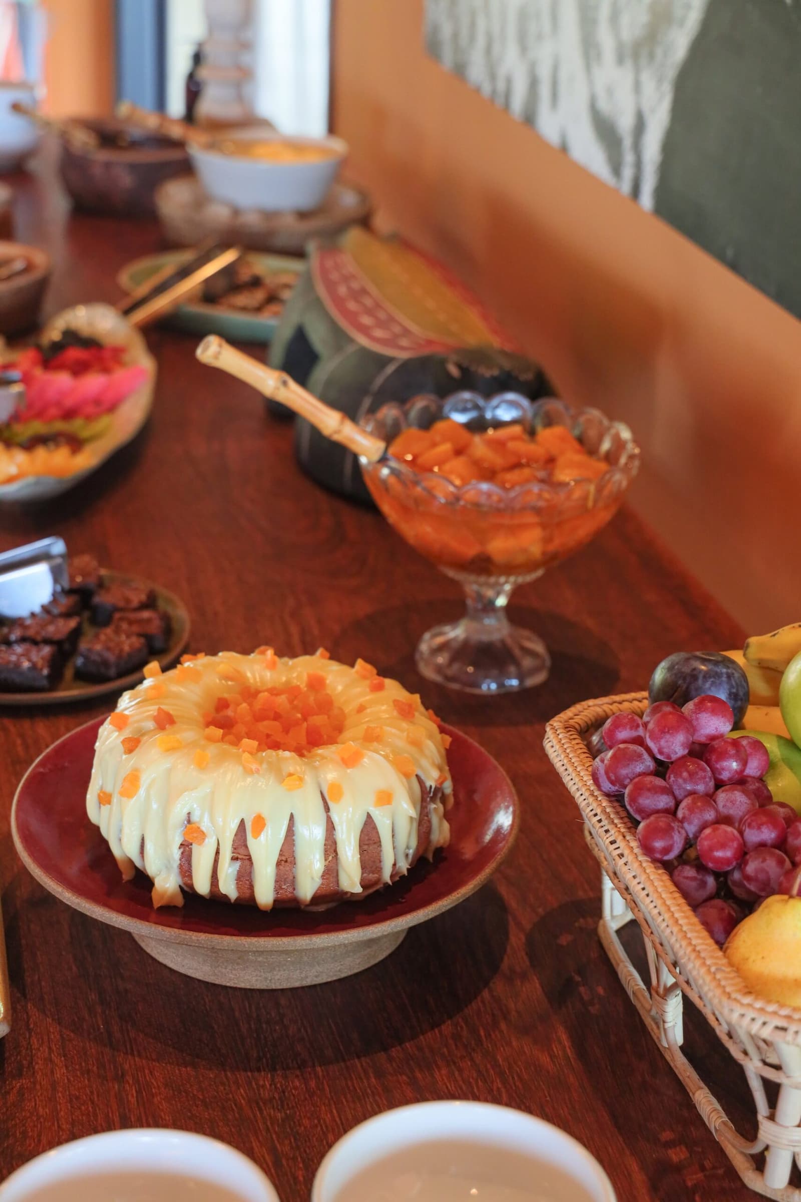 Cake before capybaras. No one ever complained. Photo: thiagolbrito A glazed bundt cake topped with diced papaya sits on a red ceramic stand on a wooden buffet table, surrounded by a glass bowl of cubed orange fruit, a basket of red grapes and fresh fruit, brownies, and mixed tropical fruit bowls — part of the generous breakfast spread on a luxury Pantanal and Amazon trip