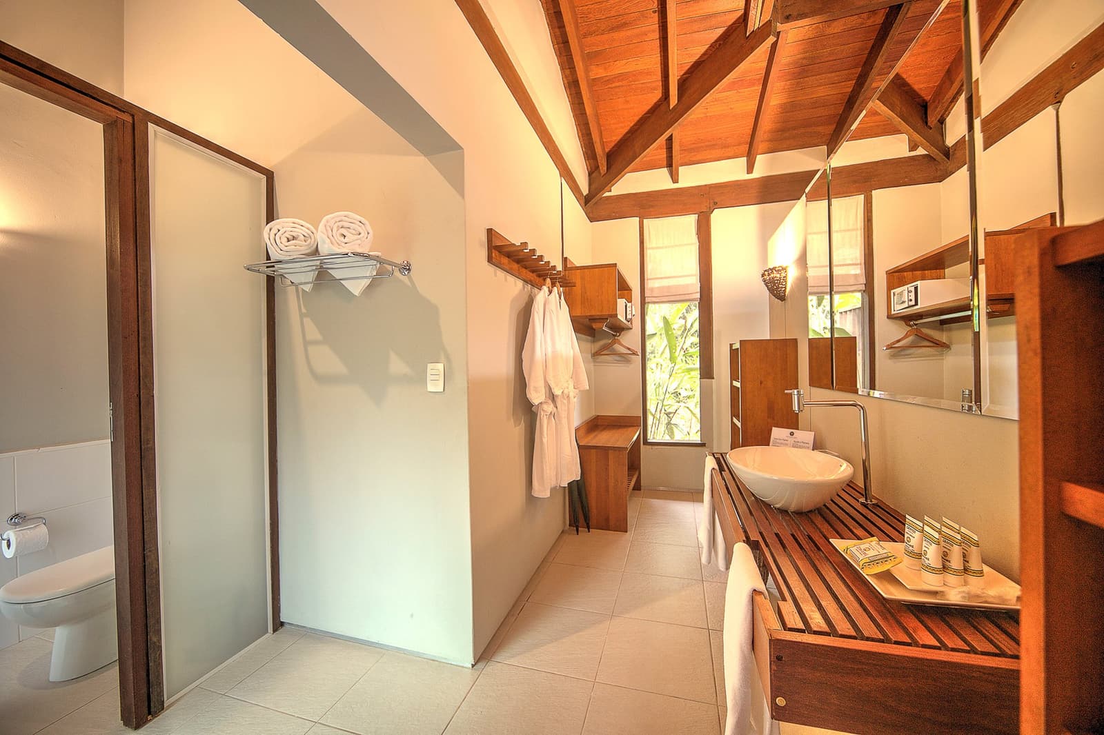 Crisp towels, a timber ceiling, and greenery just past the glass. Roughing it, this is not. Photo: | SamuelMelim Photography Spacious lodge bathroom with exposed timber beam ceiling, a vessel sink on a slatted hardwood vanity, folded white towels, bathrobes, and a window overlooking green foliage — part of a luxury Pantanal and Amazon trip stay experience