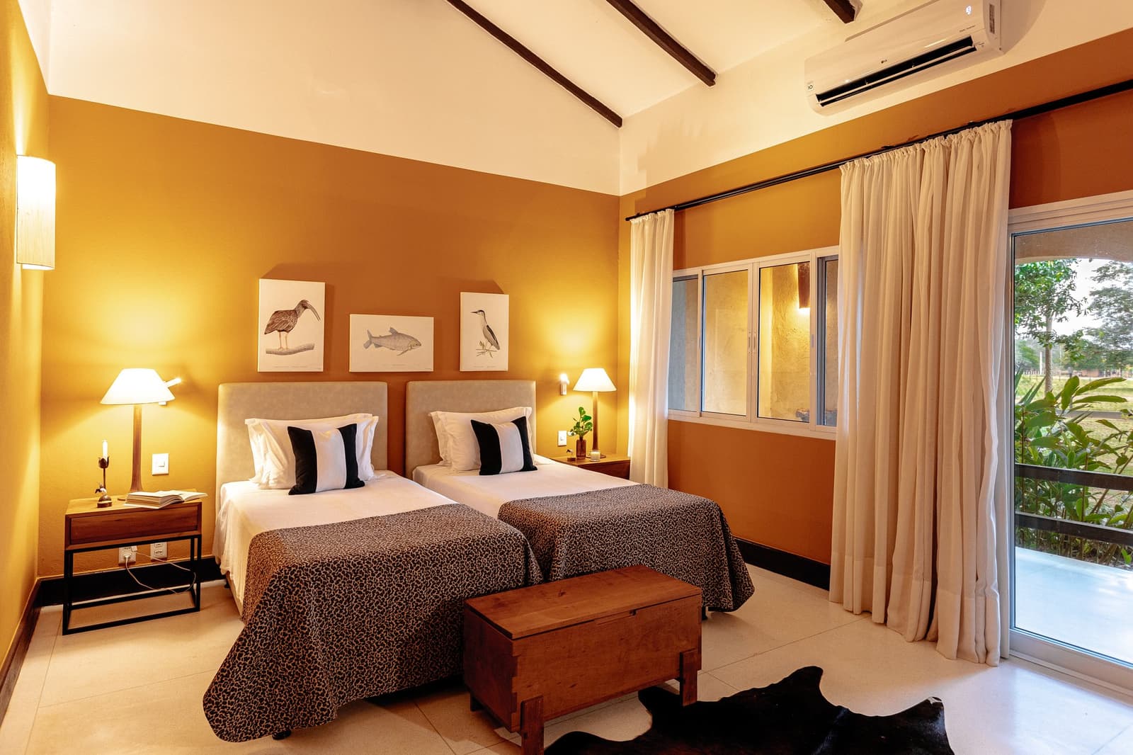 Animal art on the walls, real animals just outside the door. Photo: Felipe Castellari Twin-bedded room on a luxury Pantanal and Amazon trip, with warm ochre walls, leopard-print bedspreads, a rustic wooden chest, and botanical wildlife illustrations above the headboards