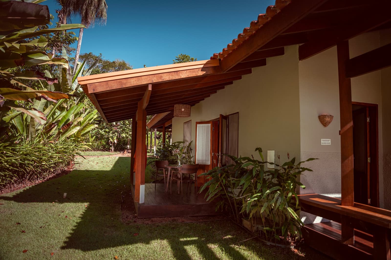 Shade, a table set for two, and the jungle starting right at the lawn's edge. A tiled-roof bungalow on a luxury Pantanal and Amazon trip, with a shaded timber veranda, wicker pendant light, outdoor dining table, and lush tropical garden surroundings under a clear blue sky