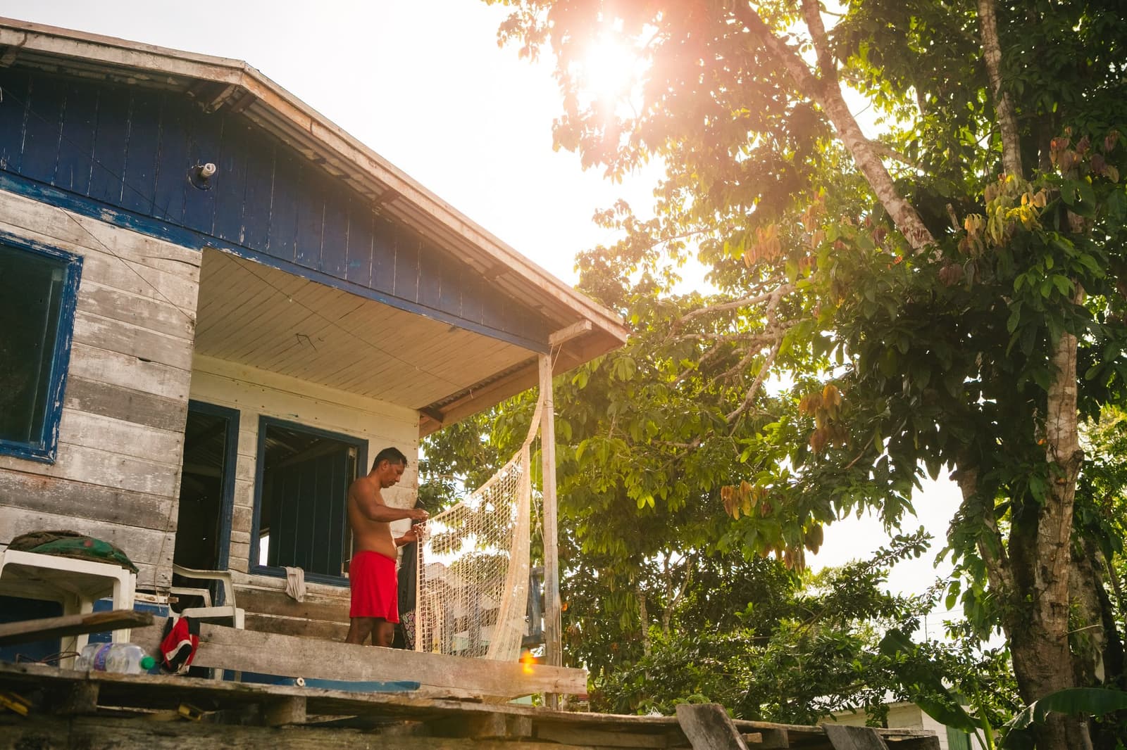 Nets don't mend themselves, and mornings here start before the forest does. Photo: Guilherme Gomes A man mending a fishing net on the porch of a weathered blue-and-grey wooden house beside the forest, warm backlit sunlight filtering through the canopy above, part of an amazon rainforest ecolodge trip in brazil that includes a riverside community visit