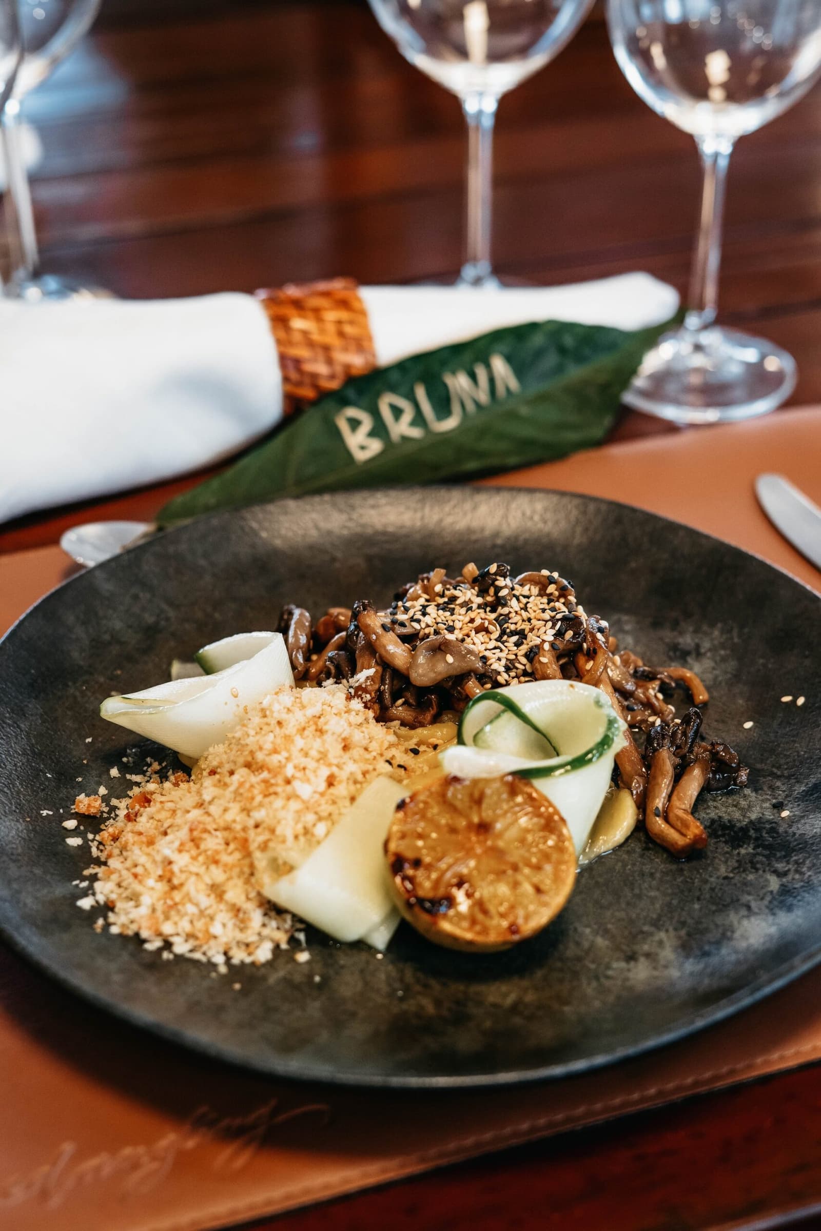 Even the place cards grow on trees out here. Sautéed mixed mushrooms with sesame seeds, shaved vegetables, a grilled round slice, and toasted breadcrumbs on a dark ceramic plate, served on a luxury Amazon cruise in Brazil with a personalized leaf place card reading 'Bruna'
