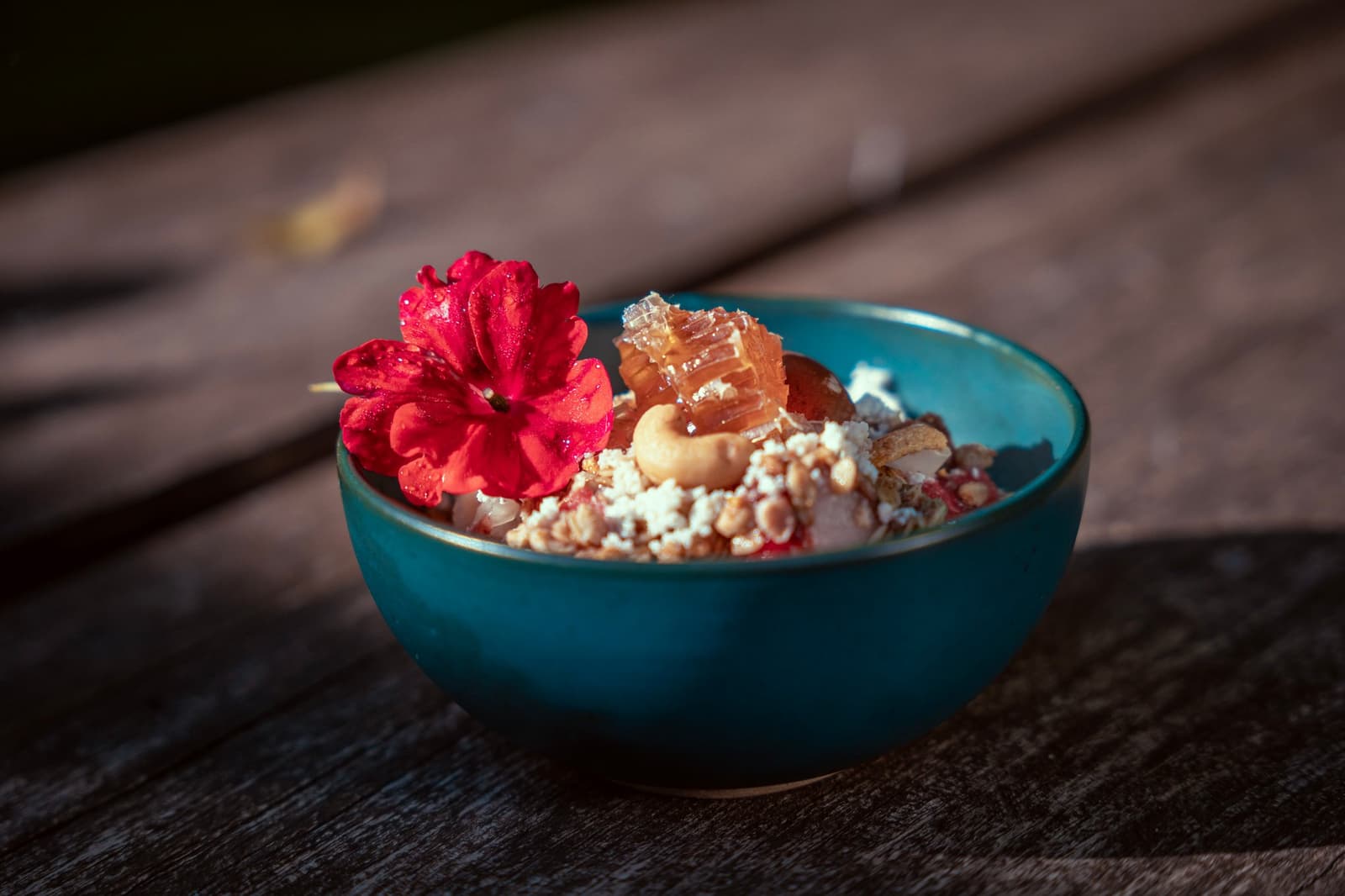 Breakfast that takes its time, honeycomb, nuts, and a flower just because. A teal ceramic bowl on a weathered wooden table, filled with granola, nuts, crumbled white cheese, and a chunk of raw honeycomb, garnished with a vivid red edible flower — part of the lodge breakfast on a luxury Pantanal and Amazon trip