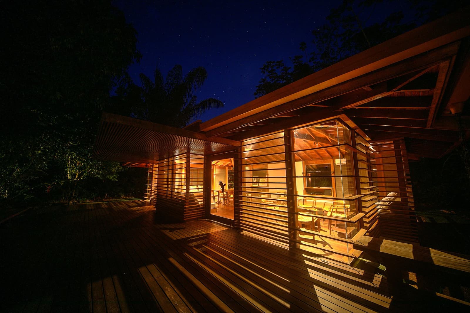 Dinner done, stars out, forest humming, the lodge earns its keep after dark. Photo: Samuel Melim A timber-and-glass jungle lodge glows warm amber at night on a luxury pantanal and amazon trip, with a wide hardwood deck, louvred walls, and a star-flecked deep-blue sky above the surrounding forest canopy