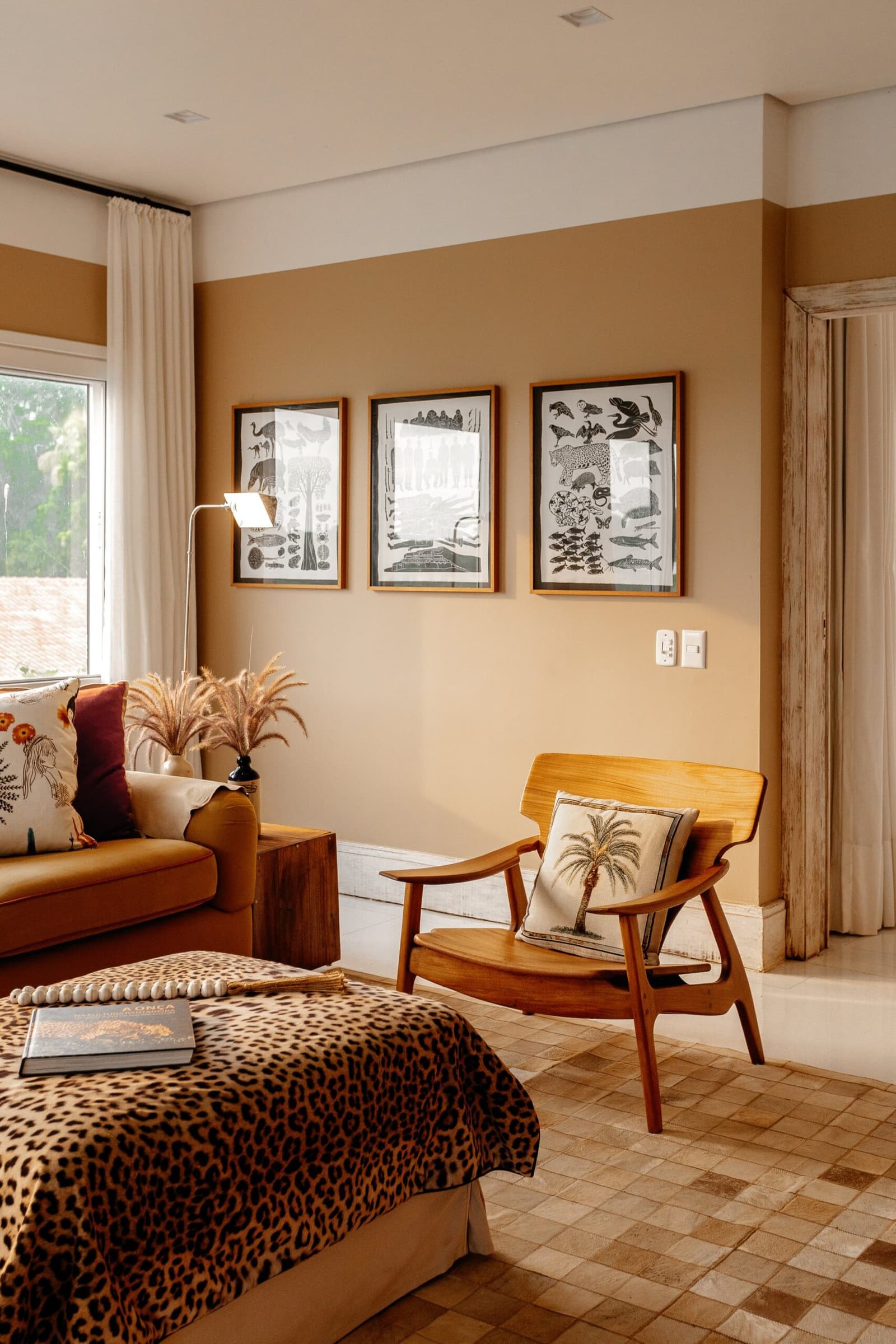 Nature does not stay outside, it follows you right into your room. Photo: Felipe Castellari Lodge suite interior on a luxury Pantanal and Amazon trip, featuring a leopard-print bedspread, mid-century wooden armchair with a palm-tree cushion, three framed wildlife illustration prints on a warm ochre wall, and a cowhide-patterned tile floor