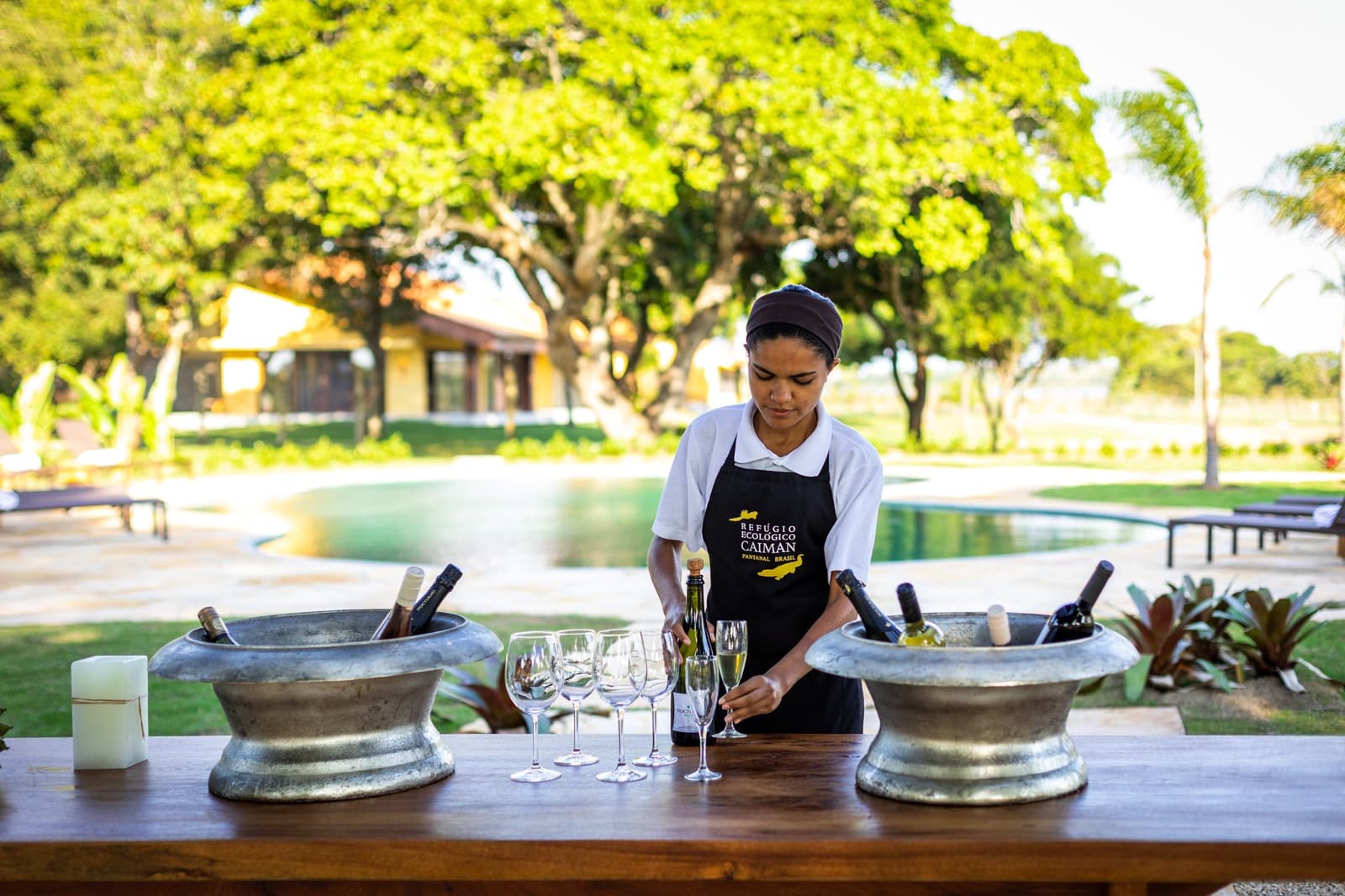 Cold bottles, crystal glasses, and someone who actually knows how to set a bar. A Refúgio Ecológico Caiman staff member in a branded apron arranges champagne flutes and wine bottles chilled in pewter buckets on a wooden bar, with the lodge pool and garden visible behind her on this luxury Pantanal and Amazon trip
