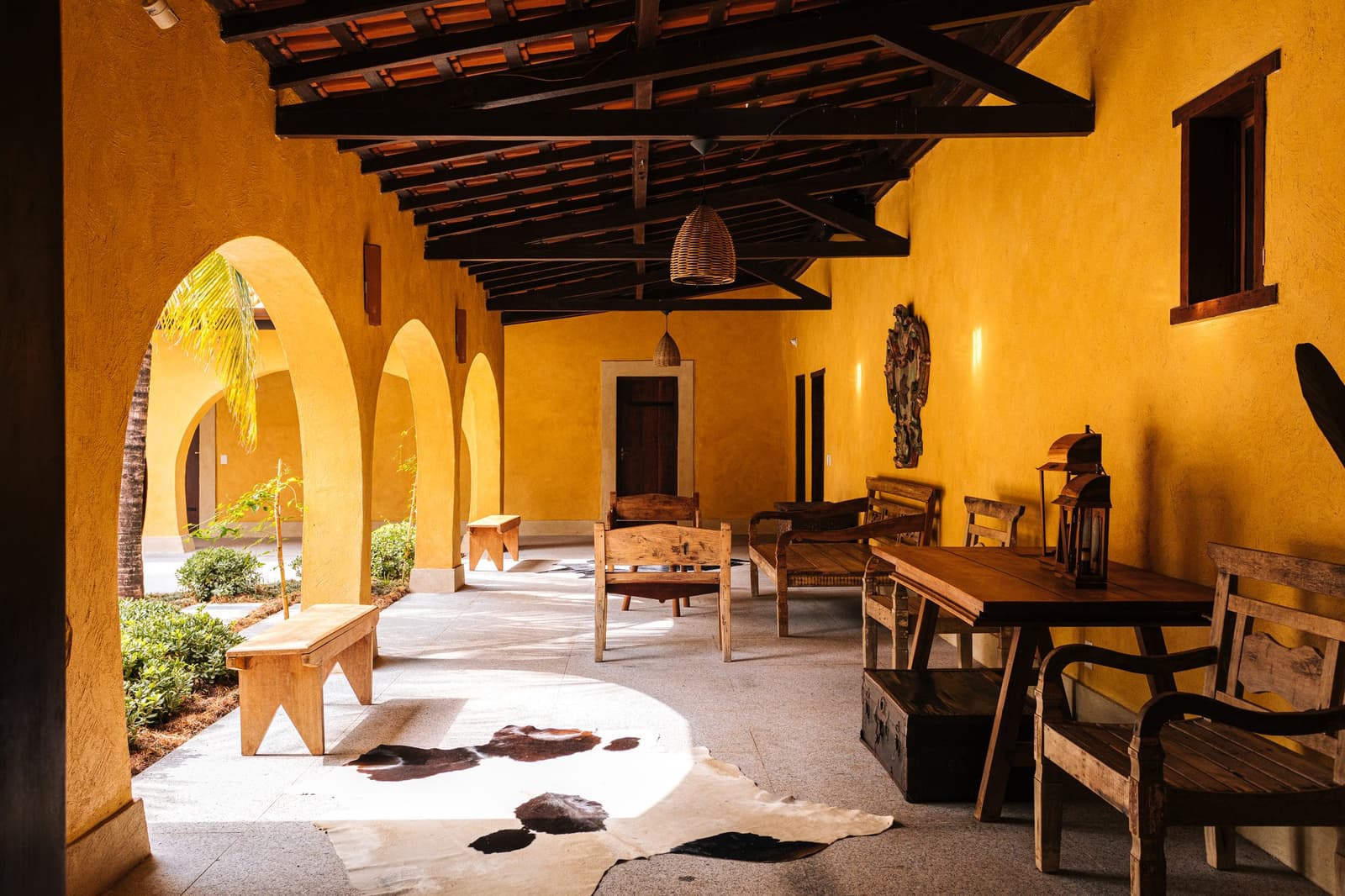 Arches, timber beams, and a cowhide rug, the Pantanal has its own idea of comfort. Covered veranda of a luxury pantanal and amazon trip lodge, with golden-yellow arched walls, dark timber ceiling beams, rustic hardwood furniture, wicker pendant lights, and a cowhide rug on the stone floor