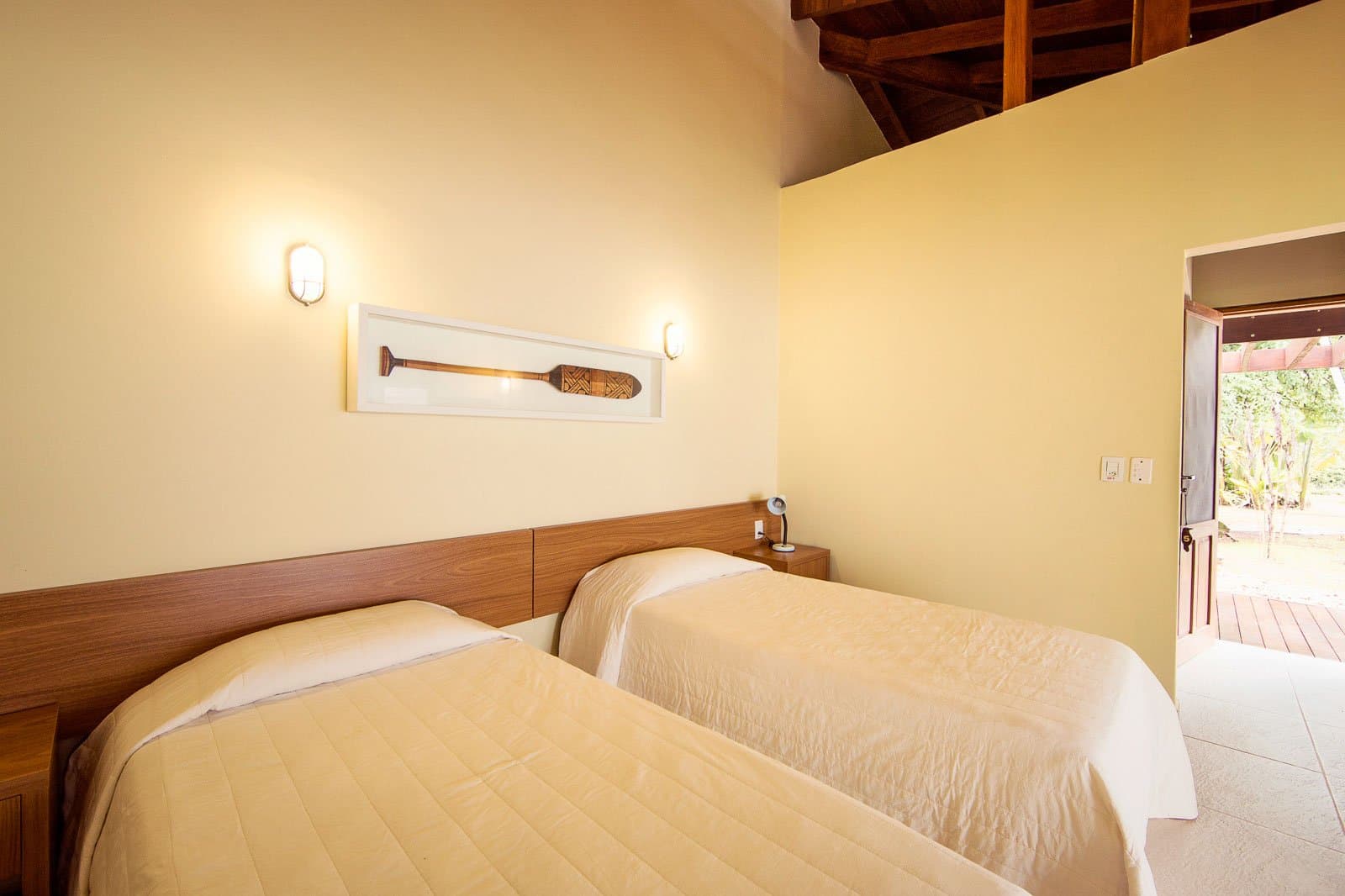 Two beds, a carved paddle, and the forest just past that open door. Photo: | SamuelMelim Photography Twin-bed room at a luxury Pantanal and Amazon trip lodge, with cream quilted bedspreads, a warm wooden headboard, wall sconces, a framed decorative paddle on the wall, exposed timber ceiling beams, and an open door leading to a sunlit outdoor deck