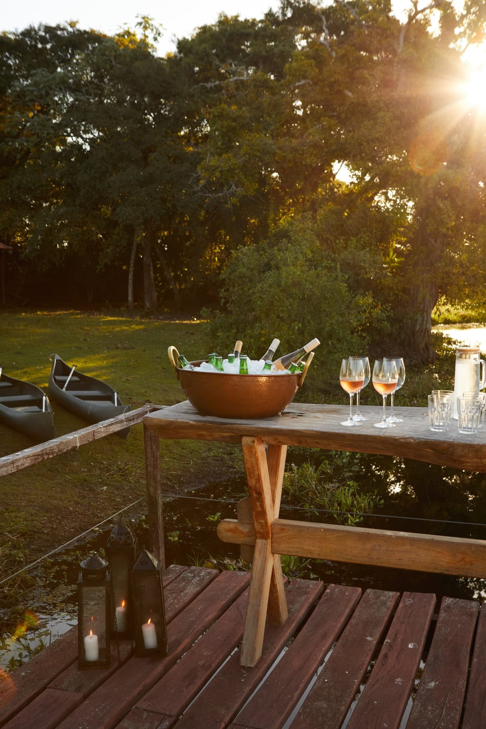 Sundowners sorted before the canoes are even put away. A copper ice bucket filled with chilled bottles and rosé wine glasses set on a rustic wooden table on a riverside deck at golden hour, with canoes on the grass and candles glowing below, on a luxury Pantanal and Amazon trip