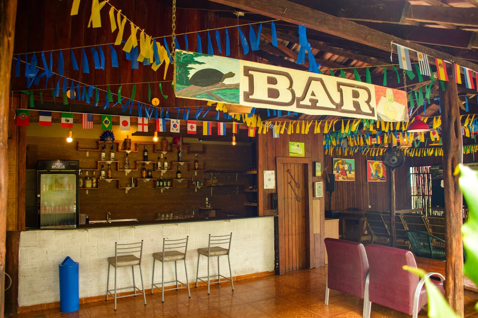 Flags from a dozen countries, one hand-painted turtle, and a cold beer fridge. The bar earns its welcome. Rustic open-air bar at a jungle lodge on an amazon and pantanal tour, decorated with international flags, blue and yellow bunting, and a hand-painted sign featuring a turtle above the word BAR, with bottles on wooden shelves and bar stools along the counter