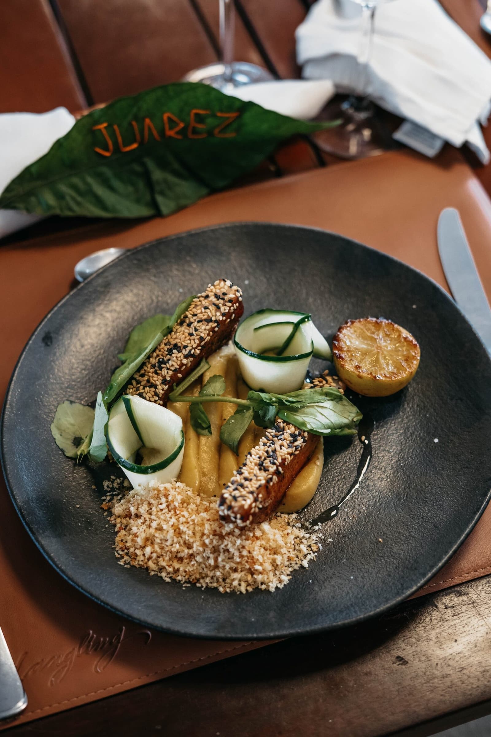 Sesame crust, cucumber spirals, charred citrus. The kitchen is clearly having fun. Sesame-crusted protein fingers with golden purée, cucumber rolls, toasted breadcrumbs, and a charred citrus half on a dark plate at Juarez restaurant, part of the luxury Amazon cruise in Brazil dining experience