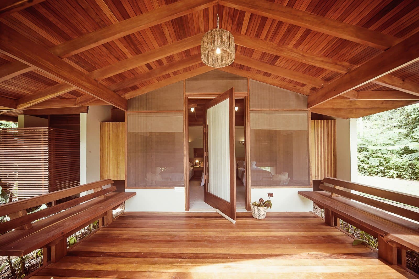 Open the doors, settle on the bench, let the forest do the rest. Photo: | SamuelMelim Photography Wooden veranda of a jungle lodge cabin on a luxury Pantanal and Amazon trip, with two bench seats flanking open double doors that lead into a softly lit bedroom, surrounded by dense forest greenery