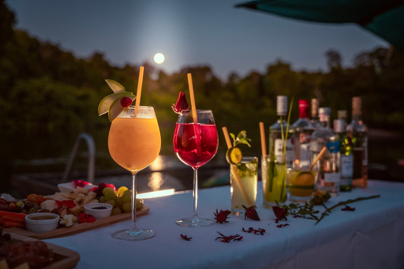 Full moon, four cocktails, and a charcuterie board. The evening just sorted itself out. Four garnished cocktails and a gratuity board on a white-draped table at dusk, with a full moon rising over the treeline on a luxury Pantanal and Amazon trip