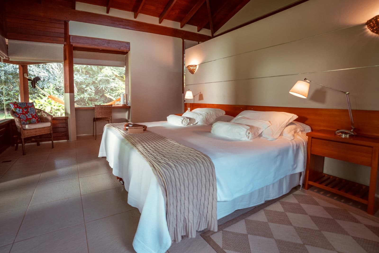 Crisp white sheets, warm wood, and the forest right there at the window. Spacious lodge bedroom on a luxury Pantanal and Amazon trip, with a king bed dressed in white linens, warm timber ceiling beams, bedside lamps, and large windows opening onto dense green forest
