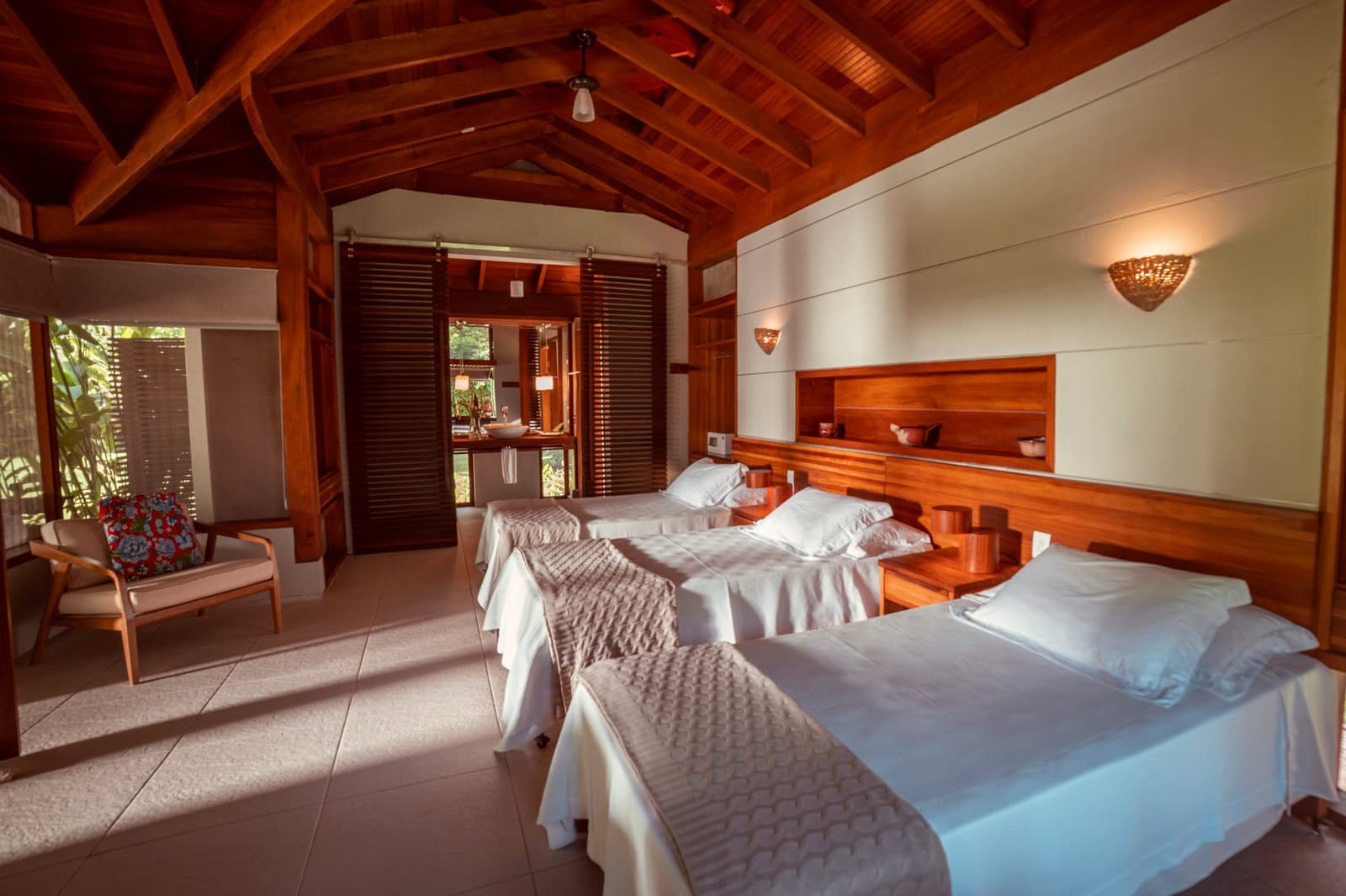 Quiet, well-lit, and built from wood that still smells like forest. Photo: Samuel Melim Twin-bed guest room on a luxury Pantanal and Amazon trip, featuring a soaring timber-beam ceiling, warm wood headboard with built-in shelving, woven wall sconces, and a sliding louvred door opening to an en-suite bathroom, with tropical garden views through slatted windows