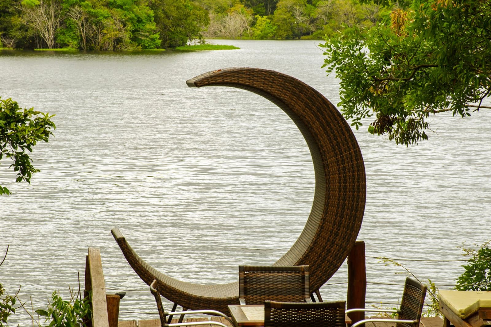 That chair was designed by someone who never wanted to leave the riverbank. A crescent-shaped wicker lounge chair on a wooden deck overlooking a calm river lined with dense green forest, part of an amazon and pantanal tour lodge stay