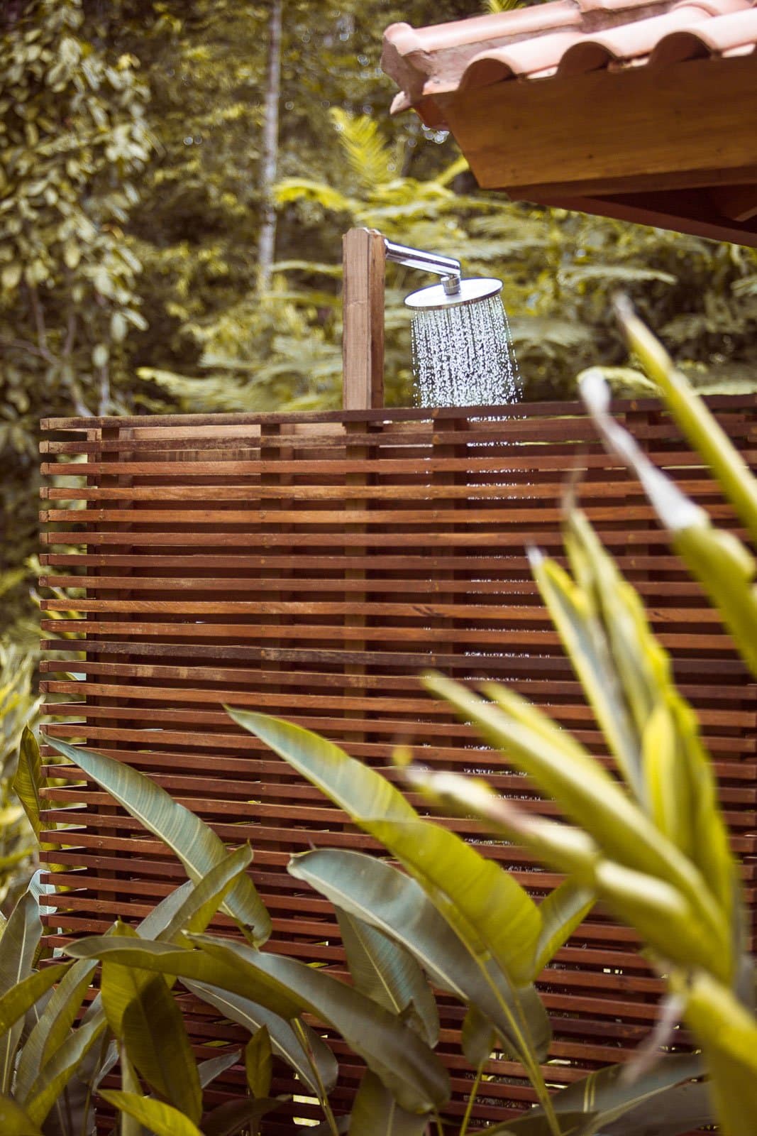 Rinse off in the forest, birds optional but likely. Photo: | SamuelMelim Photography An open-air rain shower head running water above a slatted hardwood privacy screen, surrounded by lush tropical foliage and a terracotta-tiled roof — lodge amenity on a luxury Pantanal and Amazon trip