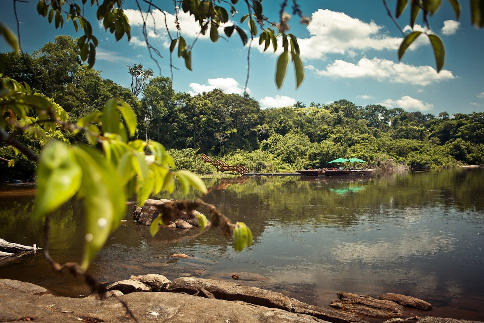 Rock, river, and a shaded deck just far enough into the trees to feel earned. Photo: Samuel Melim (c) A calm forest river reflects green canopy and blue sky, with a wooden riverside lodge platform shaded by teal umbrellas visible through overhanging leaves — part of a luxury Pantanal and Amazon trip experience deep in the rainforest.