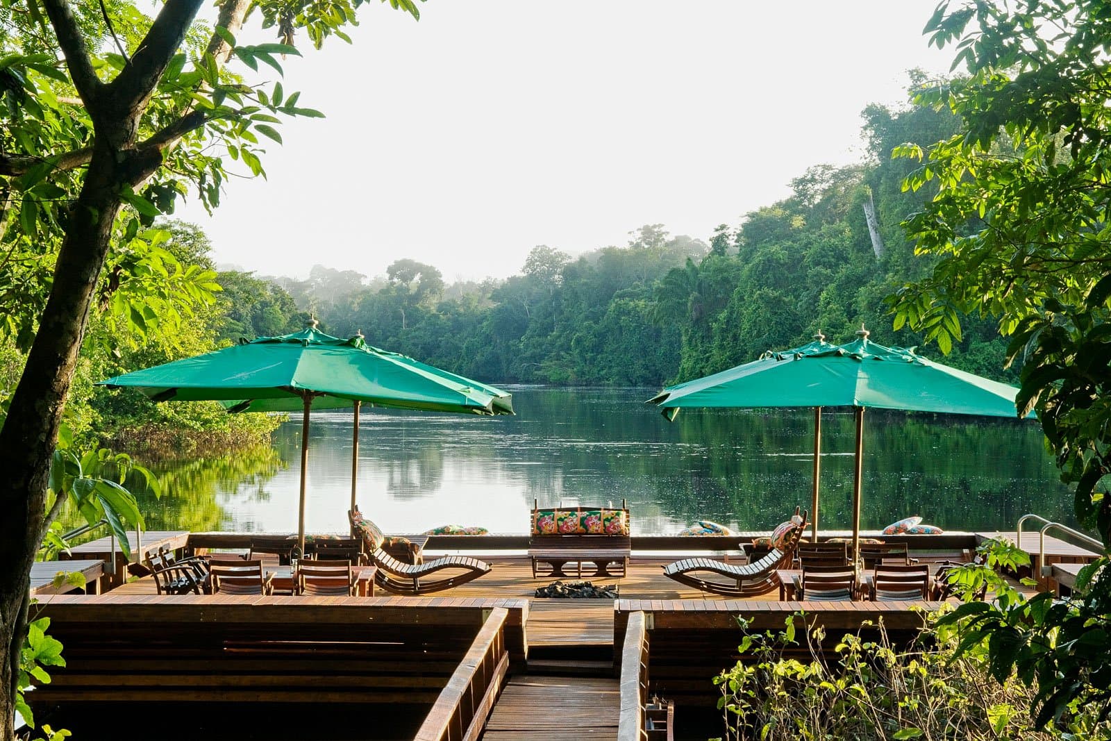 The forest comes right to the edge, and the river does the rest. Photo: Luis Gomes A hardwood deck with wooden sun loungers and green parasols overlooks a calm river flanked by dense rainforest canopy on a luxury Pantanal and Amazon trip