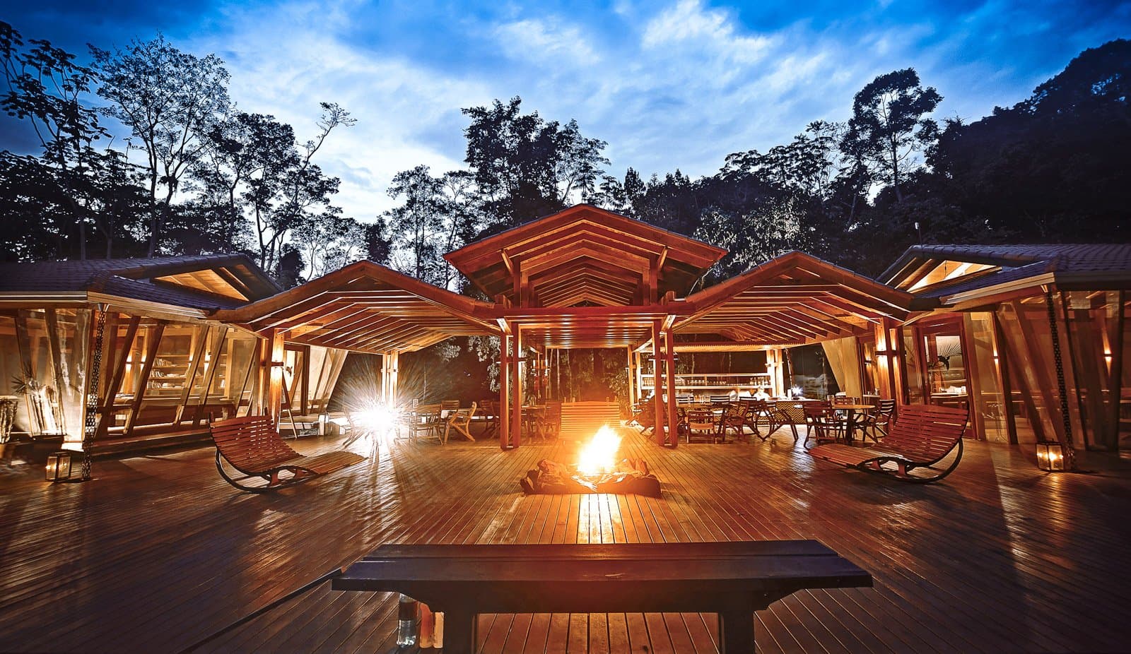 Drinks, a fire, and the forest deciding when the evening ends. Photo: Samuel Melim (c) Open-air timber lodge deck at dusk on a luxury Pantanal and Amazon trip, with a campfire burning at the centre, rocking chairs to either side, a bar and dining area glowing under peaked wooden roofs, and tall forest trees silhouetted against a blue twilight sky