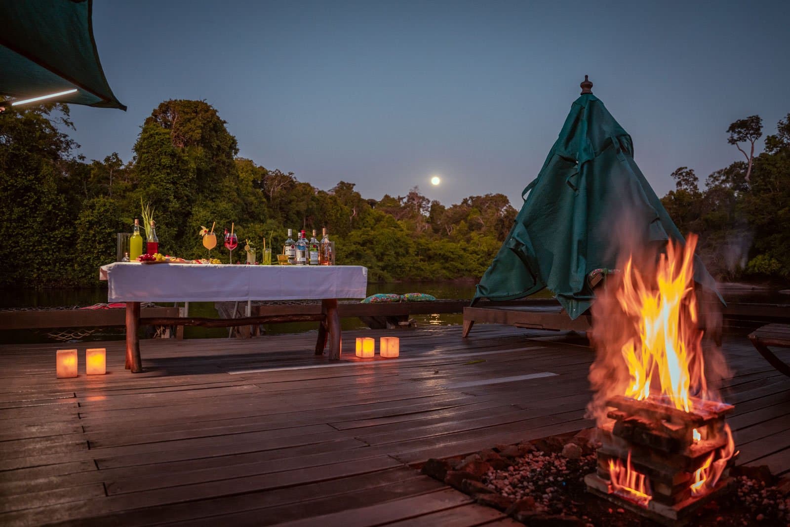 Cocktails, candlelight, and a full moon, the forest sets the mood without being asked. Photo: Samuel Melim A wood deck beside a forest river at dusk, set for a luxury Pantanal and Amazon trip evening: a crackling campfire in the foreground, paper lantern candles along the boards, and a table dressed in white linen loaded with cocktails and spirits, with a full moon rising above the treeline.