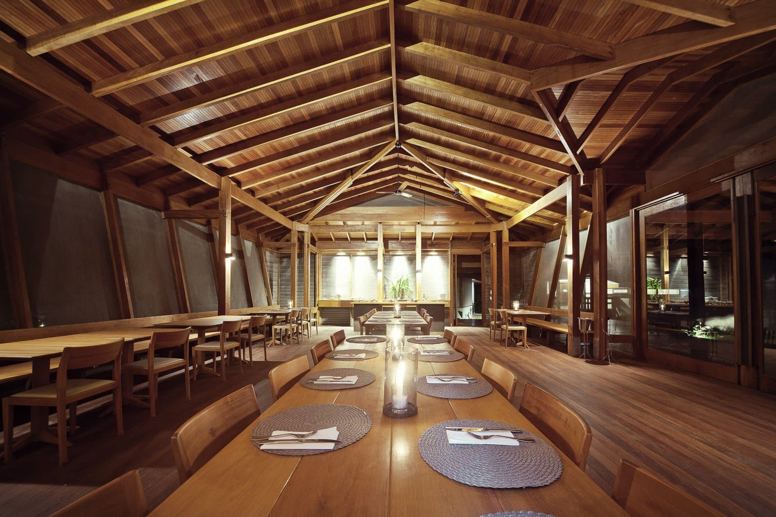 Good food, good company, and a ceiling built to impress without trying. Photo: Samuel Melim (c) Warmly lit timber dining room with vaulted wooden ceiling, long communal table set with woven placemats and candles, and smaller tables along the sides at a luxury Pantanal and Amazon trip lodge