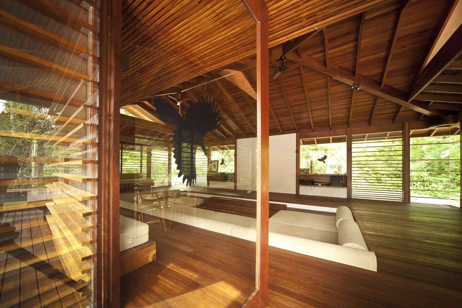 Hardwood, white cushions, forest on every side. This is where you decompress after a long day on the trail. Photo: Samuel Melim (c) Open-plan common room of a luxury pantanal and amazon trip lodge, featuring warm hardwood floors, exposed timber roof beams, white day-bed sofas, louvred shutters on three sides, and a large decorative bird sculpture suspended from the ceiling, with dense green forest visible through the open walls