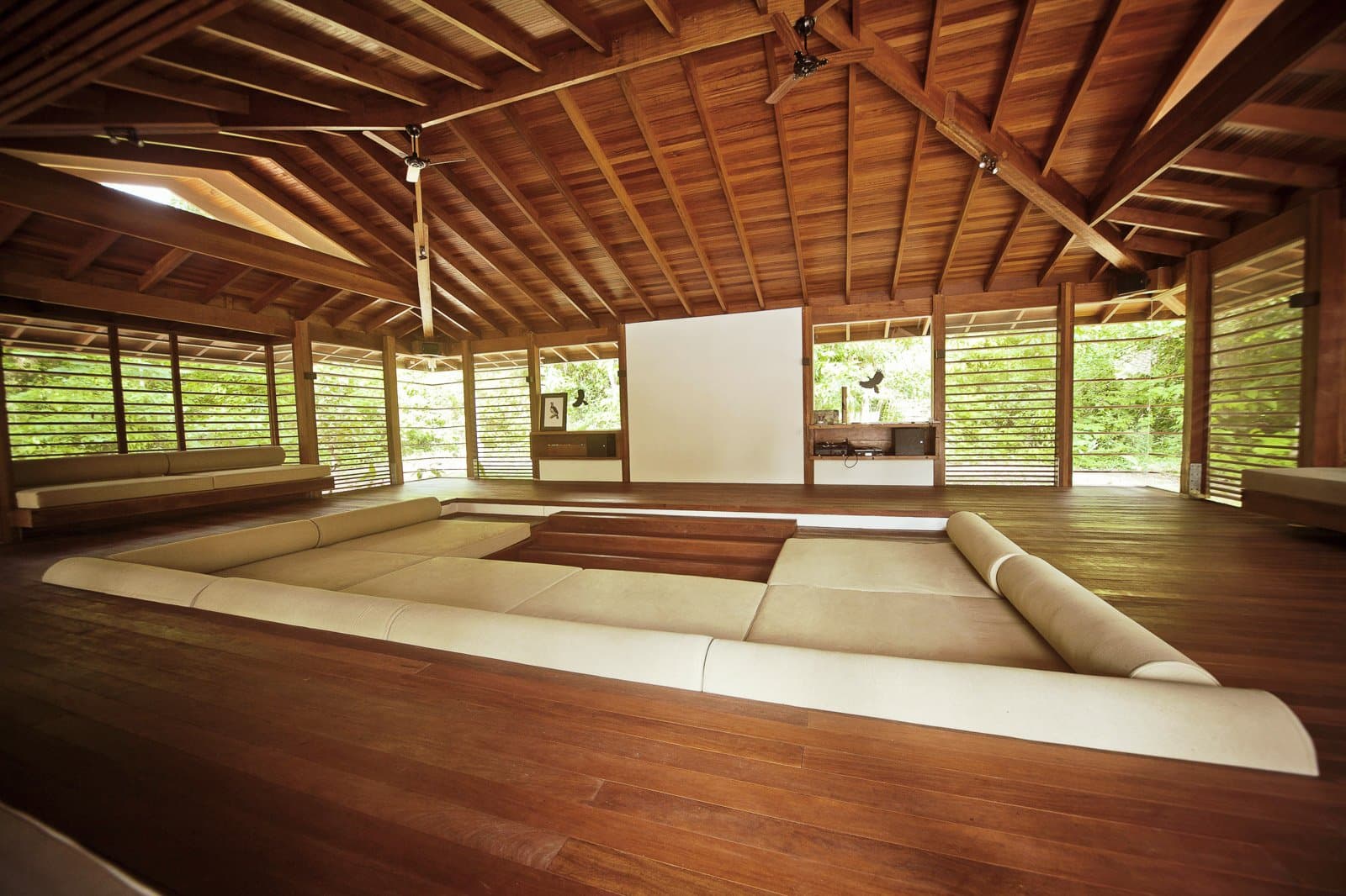 Sink into the cushions, let the forest fill the gaps in the walls. Photo: Samuel Melim (c) Open-sided timber lodge lounge on a luxury Pantanal and Amazon trip, featuring a sunken cream sectional sofa on a hardwood floor beneath a high vaulted wooden ceiling with ceiling fans, surrounded by slatted walls open to lush green forest