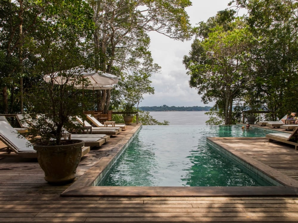 An infinity pool on a wooden deck with sun loungers overlooking a river, framed by rainforest trees at an Amazon ecolodge.