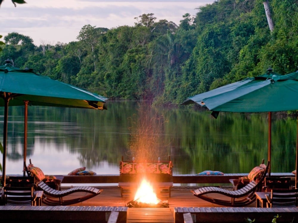 A riverside lodge deck at dusk with green umbrellas, sun loungers and a lit fire pit reflected in the calm Amazon river.