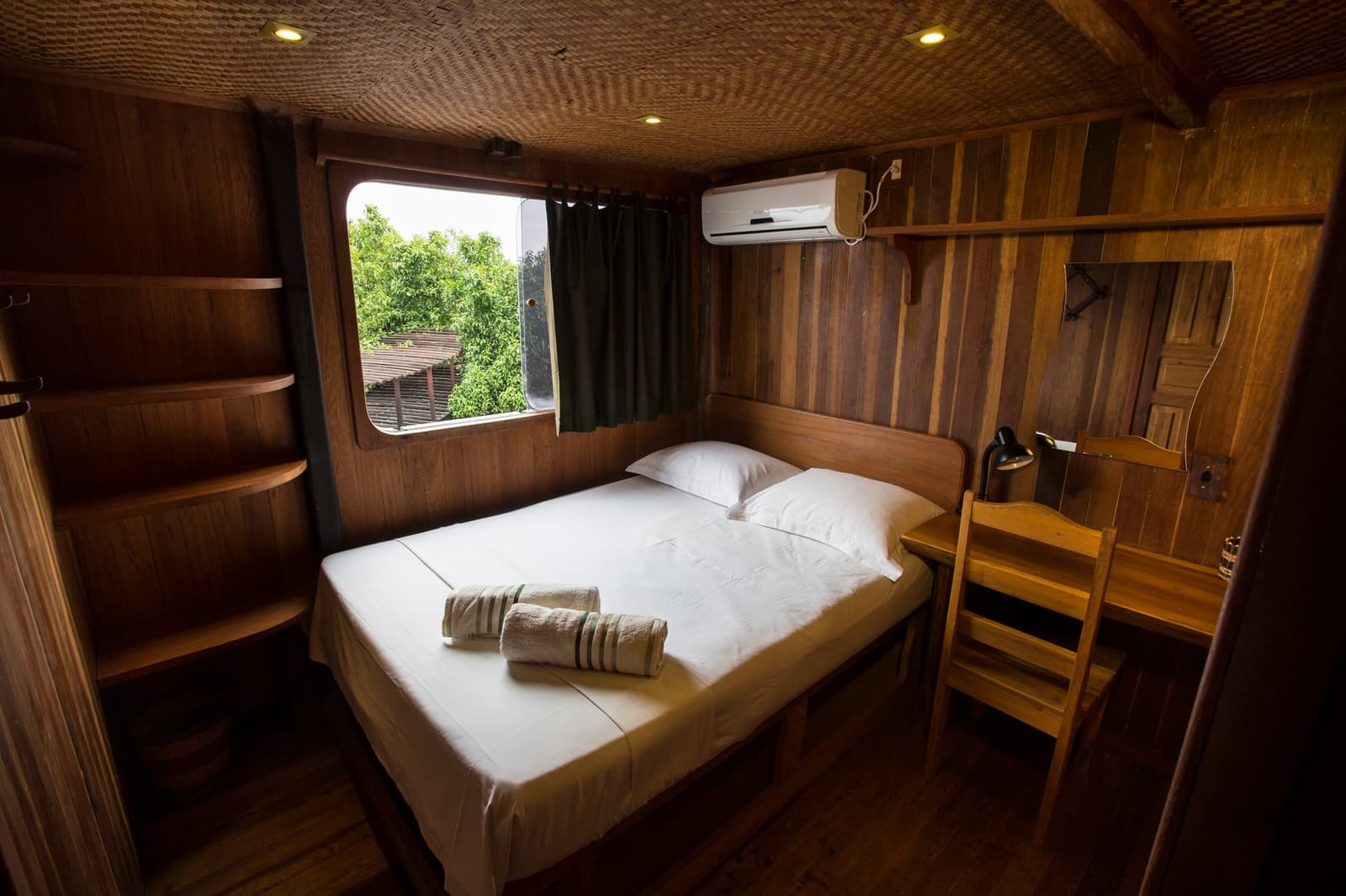 Small but considered, this cabin gives the river forest a front-row seat. Cozy wood-panelled cabin on an Amazon river cruise in Brazil, with a double bed dressed in white linen, rolled towels, open shelves, a desk, air conditioning unit, and a porthole-style window overlooking green riverside foliage