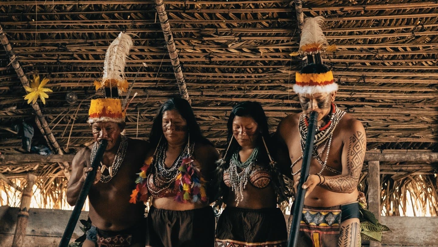 Centuries of knowledge, worn with quiet pride. Four indigenous community members in feathered headdresses, beaded necklaces, and geometric body paint perform a ceremony inside a thatched-roof maloca during the la jangada amazon cruise cultural visit