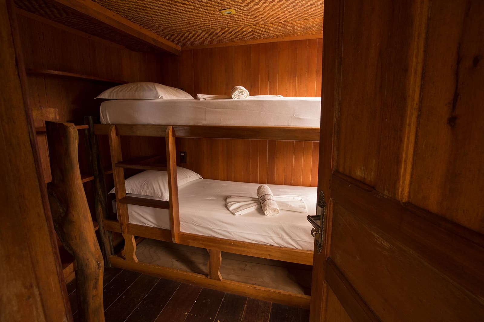 Compact, warm, and ready for you. The jungle can wait until morning. Cozy wooden bunk cabin on an amazon river cruise in Brazil, with crisp white bedding, folded towels, a rustic ladder, and a woven ceiling