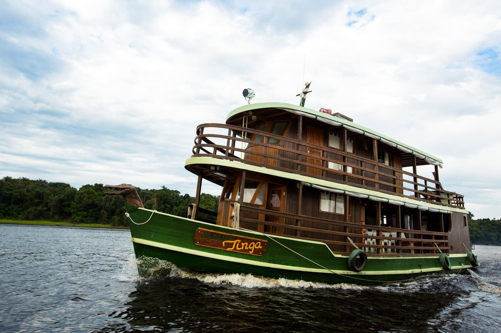 Her bow carved like a caiman, the Tinga knows exactly where she's going. Photo: sitah The Jacaré-Tinga, a three-deck wooden riverboat with a green hull, cruises a dark-water Amazon river past dense rainforest on an amazon river cruise brazil