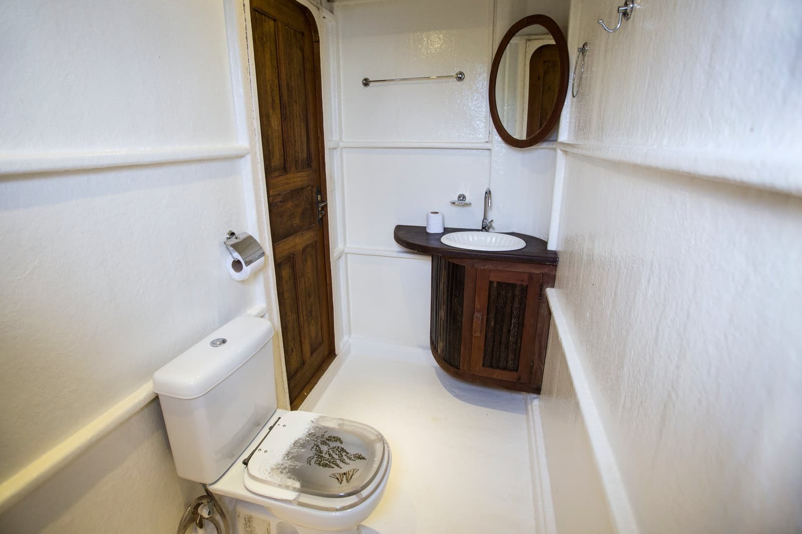 Every square metre earns its keep on a riverboat this deep in the forest. Compact private bathroom on an Amazon river cruise in Brazil, featuring a white toilet with a botanical-printed lid, a curved dark-wood vanity with round basin, oval mirror, and white-painted walls
