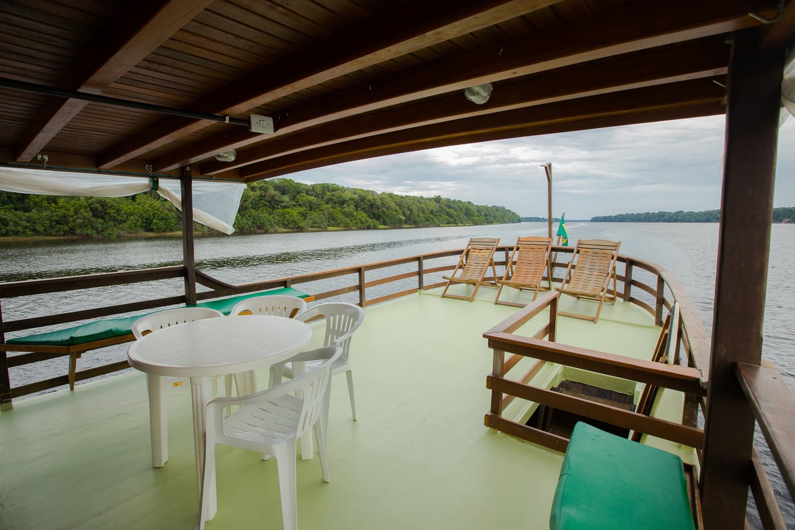 Pick a sun lounger or the shady table, the river sorts out the rest. Photo: sitah Shaded upper deck of an Amazon river cruise Brazil riverboat, with a white plastic table and chairs, wooden sun loungers, a Brazilian flag, and a calm dark-water river flanked by dense rainforest canopy stretching to the horizon.