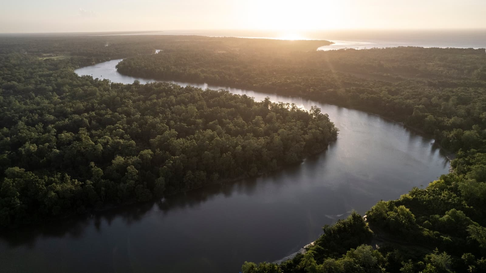 Five days, one river, and a canopy that goes on forever. Photo: Anderson Coelho Aerial view of a wide dark-water river curving through dense tropical rainforest at golden-hour sunset, with warm light glowing on the horizon where the river meets a larger body of water — the kind of landscape the la jangada amazon cruise navigates.