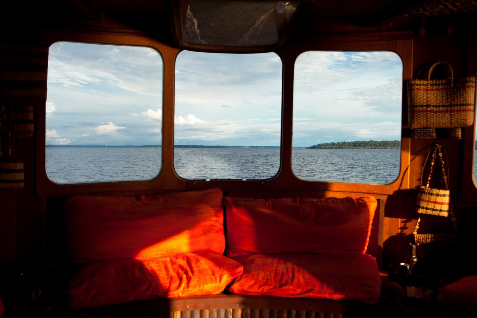 Your front-row seat to the river, no ticket required. Warm wooden interior of an Amazon river cruise Brazil riverboat, with red cushioned bench seat and three panoramic windows framing a wide river under a partly cloudy sky, woven baskets hanging on the right wall