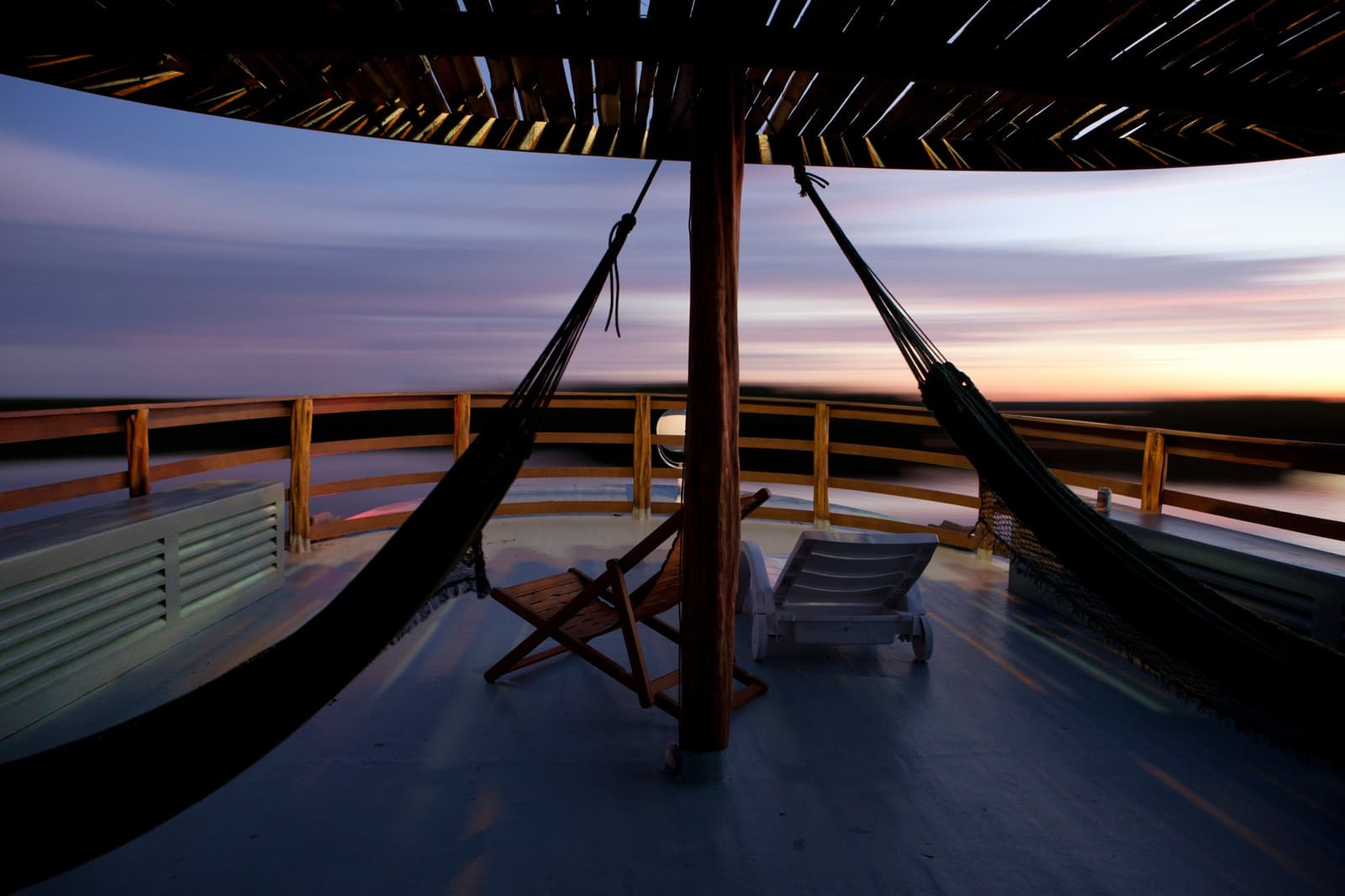 The boat keeps moving, the hammock does not care, this is how evenings are done out here. Two hammocks hang under a thatched canopy on the open upper deck of an amazon river cruise brazil riverboat, with sun loungers and a wooden folding chair framing a wide twilight horizon over a dark river floodplain