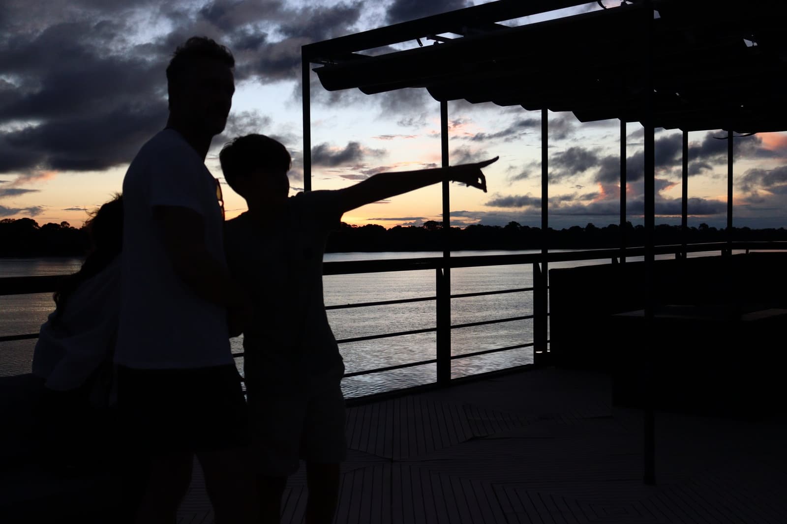 Someone always spots something first. That's what the deck is for. Two guests silhouetted on the deck of the La Jangada Amazon cruise, one pointing toward the orange and purple sunset sky over a wide river, with forested banks on the horizon