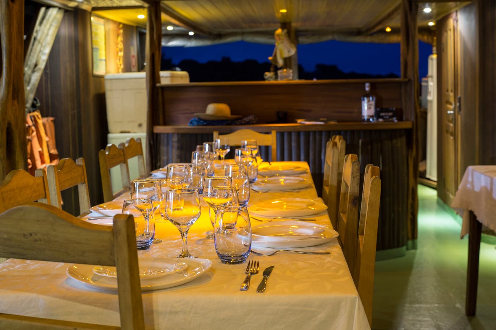 Dinner is served, the river is right there, and the night is just getting started. A long dining table set with white plates, wine glasses, and silverware glowing under warm lamplight aboard an amazon river cruise brazil riverboat, with the dark blue dusk sky visible through open windows
