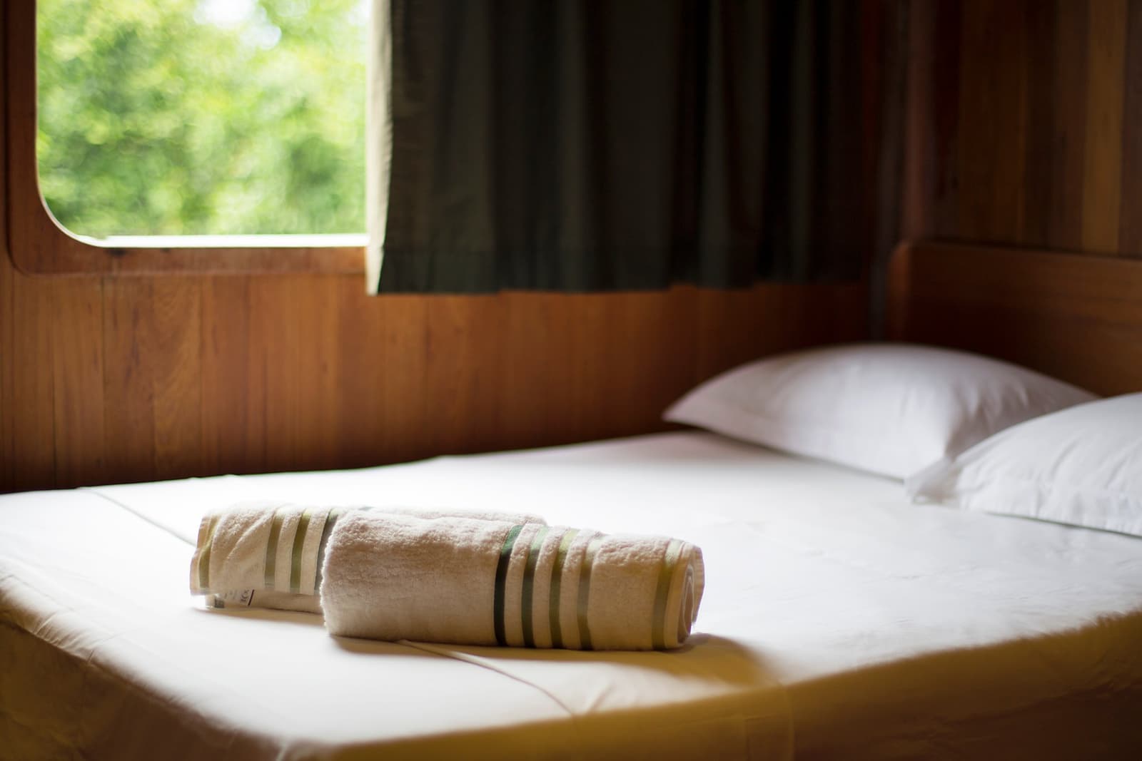 Fresh towels waiting, green canopy just outside the window. Not a bad commute. Two rolled striped towels placed on a made bed inside a wood-panelled cabin on an Amazon river cruise in Brazil, with a rounded porthole window framing green rainforest foliage outside
