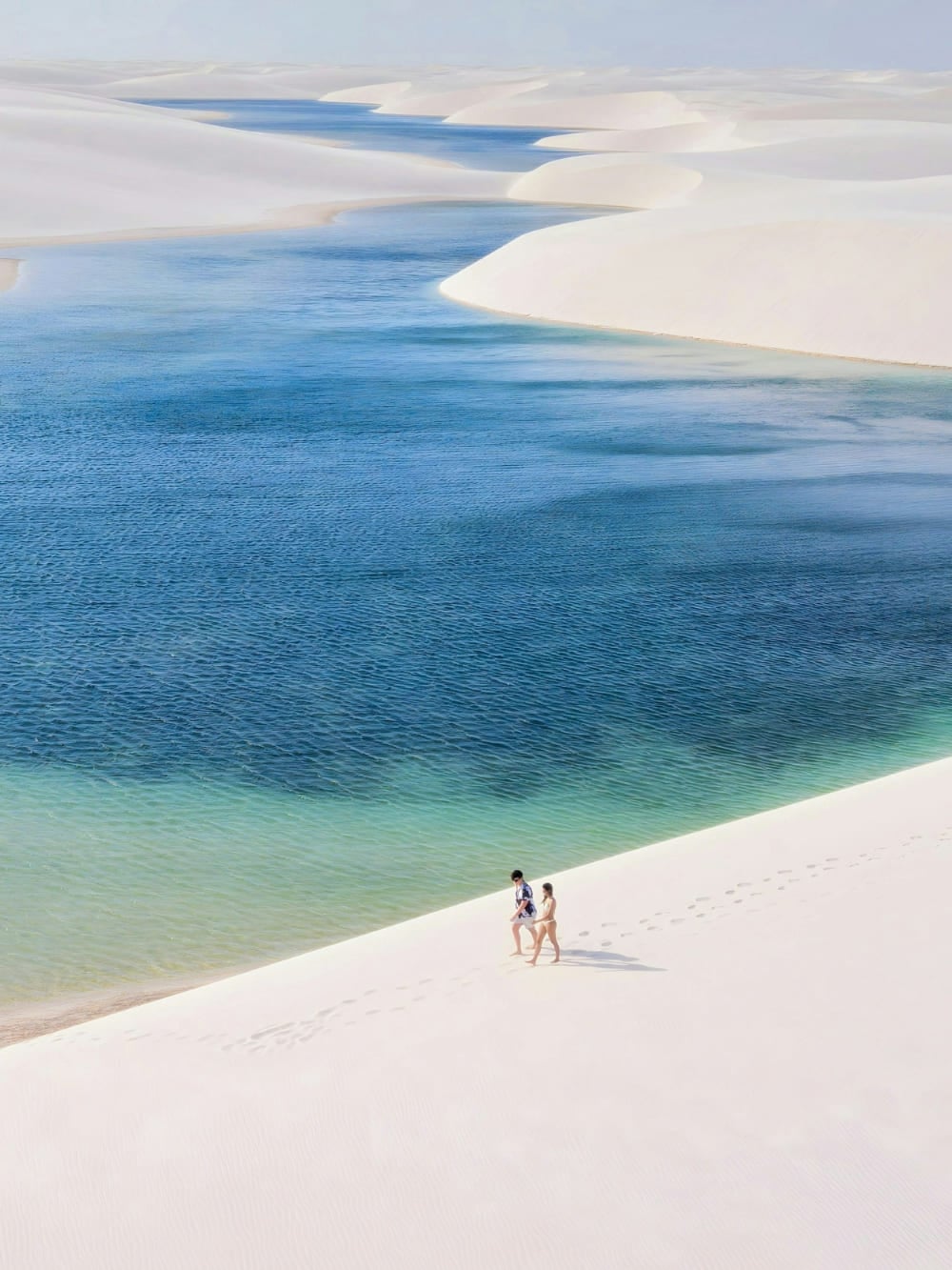 Photo: Leo Castro A couple walking along a white sand dune beside a turquoise lagoon in Lençóis Maranhenses, Brazil.