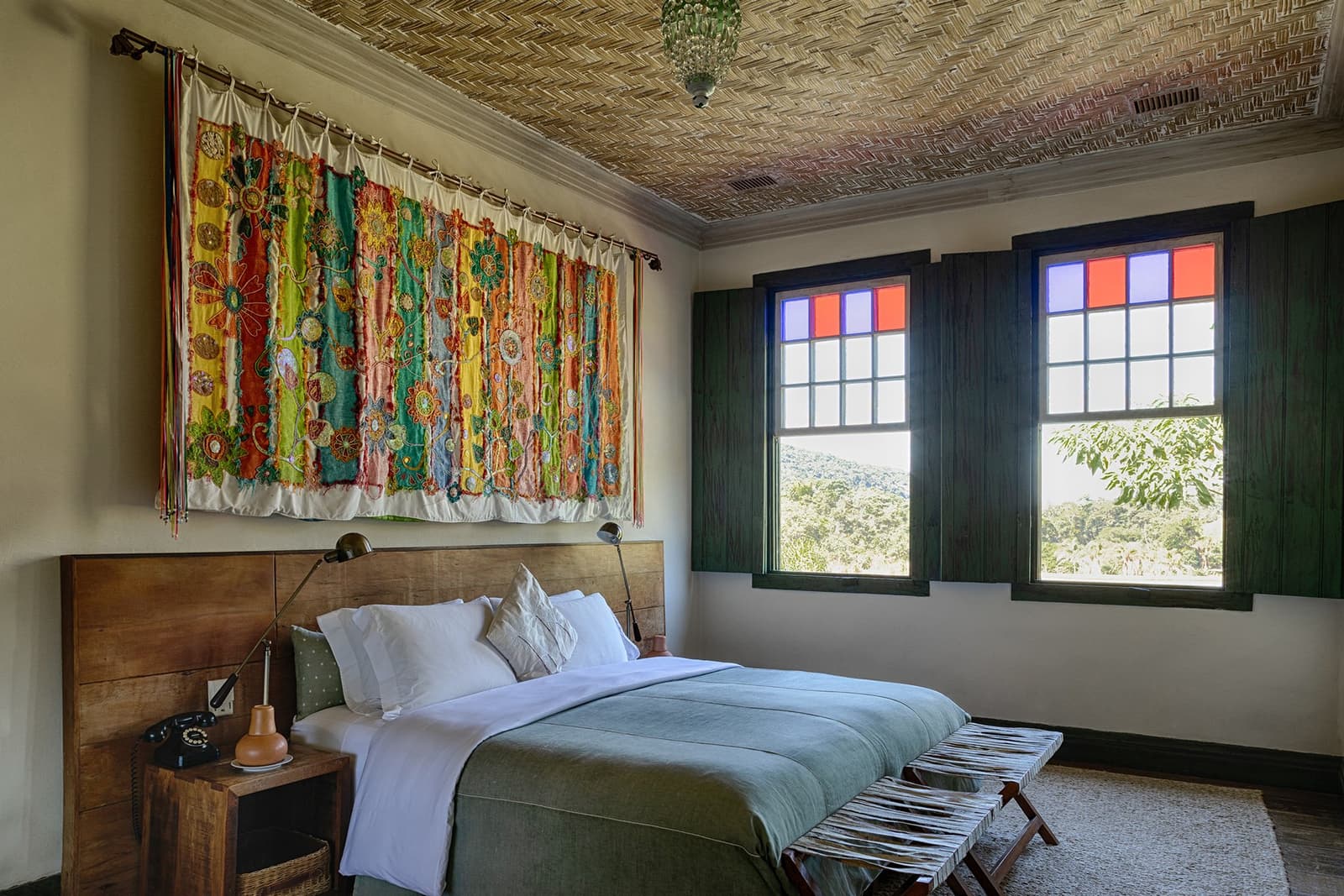 Old farmhouse bones, new linen, and a view of the forest outside your window. Photo: Henrique Mangeon A guest bedroom at the Ibiti Project's Engenho Lodge, featuring a wooden-headboard double bed dressed in white linen and a sage green throw, a large colourful embroidered textile hanging on the wall, a woven ceiling, and two colonial-style windows with red and blue stained-glass panes overlooking forested hills