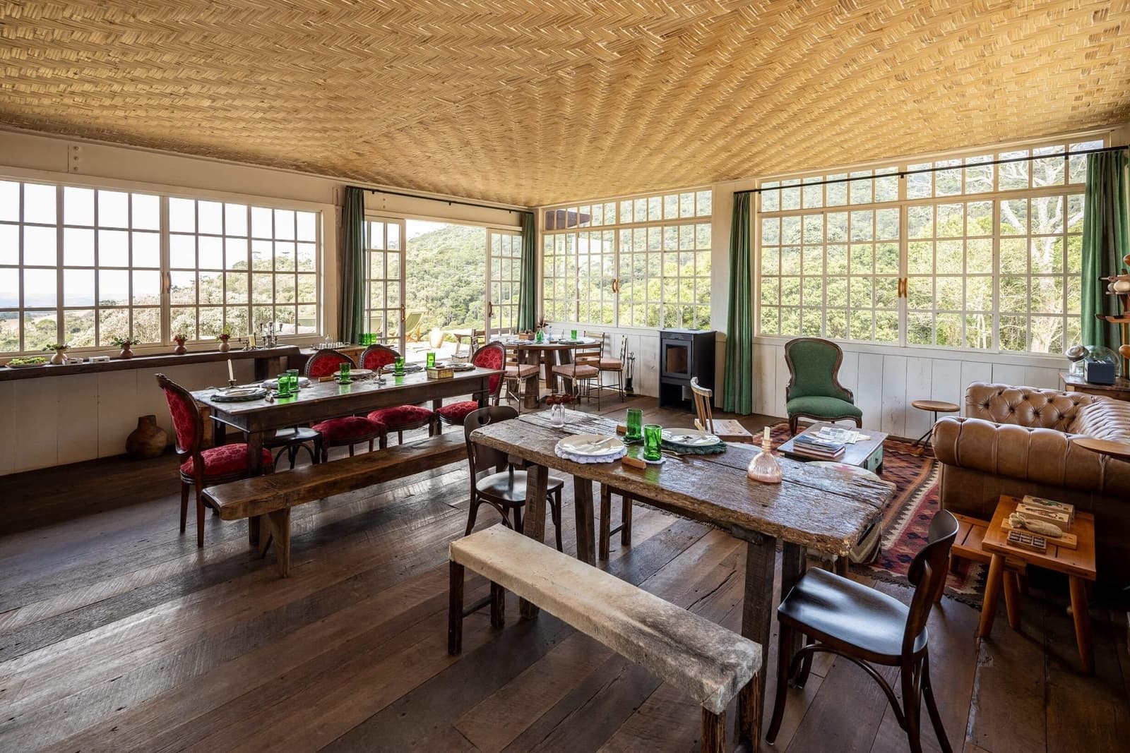 Lunch here is a proper sit-down affair, forest on every side. Rustic dining room at the Ibiti Project's Engenho Lodge, with reclaimed-wood tables, red velvet chairs, stone benches, green glassware, a woven ceiling, and floor-to-ceiling windows overlooking forest canopy