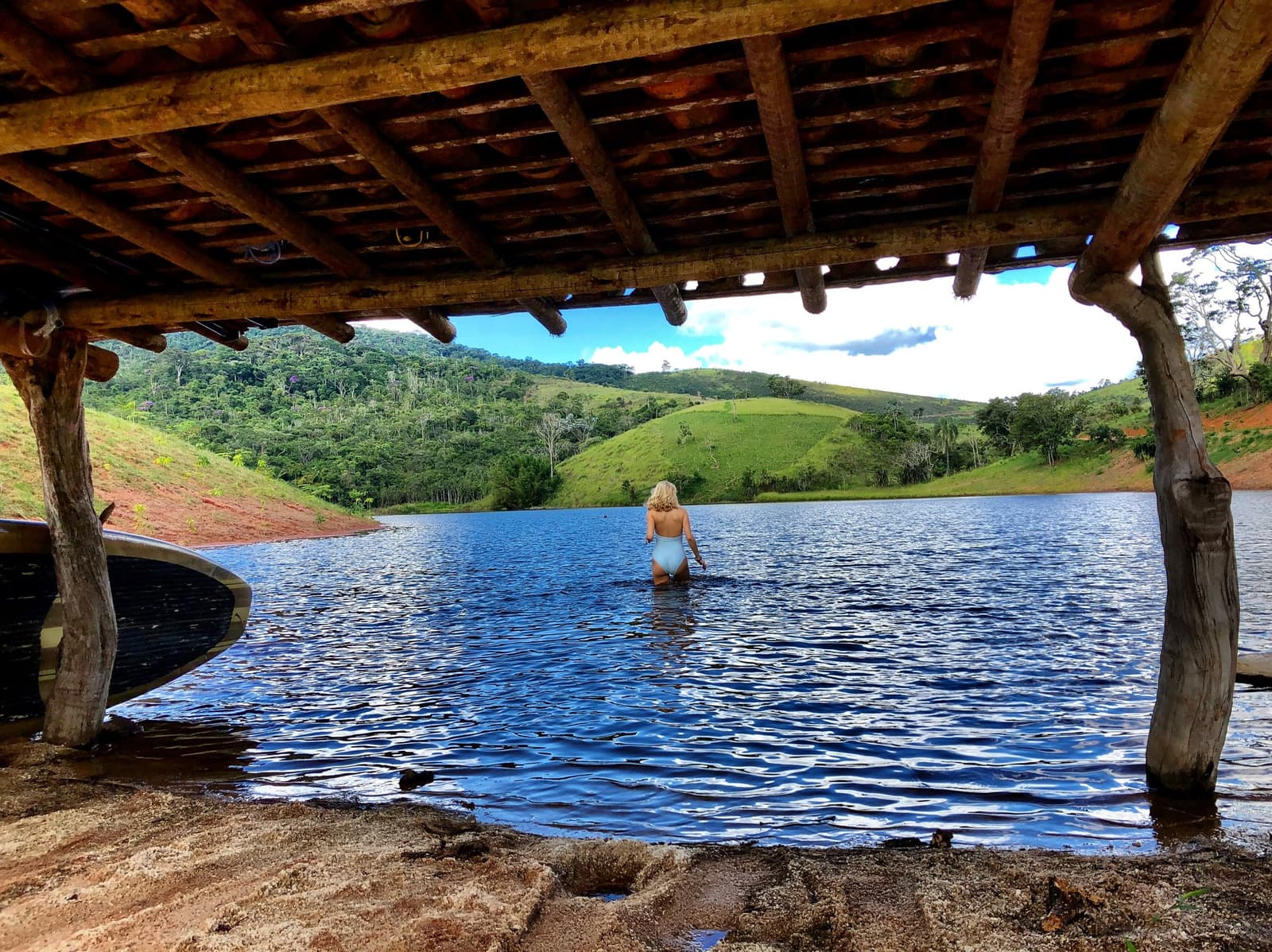 Lago Negro on a sunny afternoon. You'll want to wade in a little further. A woman in a light blue swimsuit wades into a calm lake at the Ibiti Project, framed by rustic log pillars and a wooden shelter roof, with green hills and Atlantic forest rising beyond