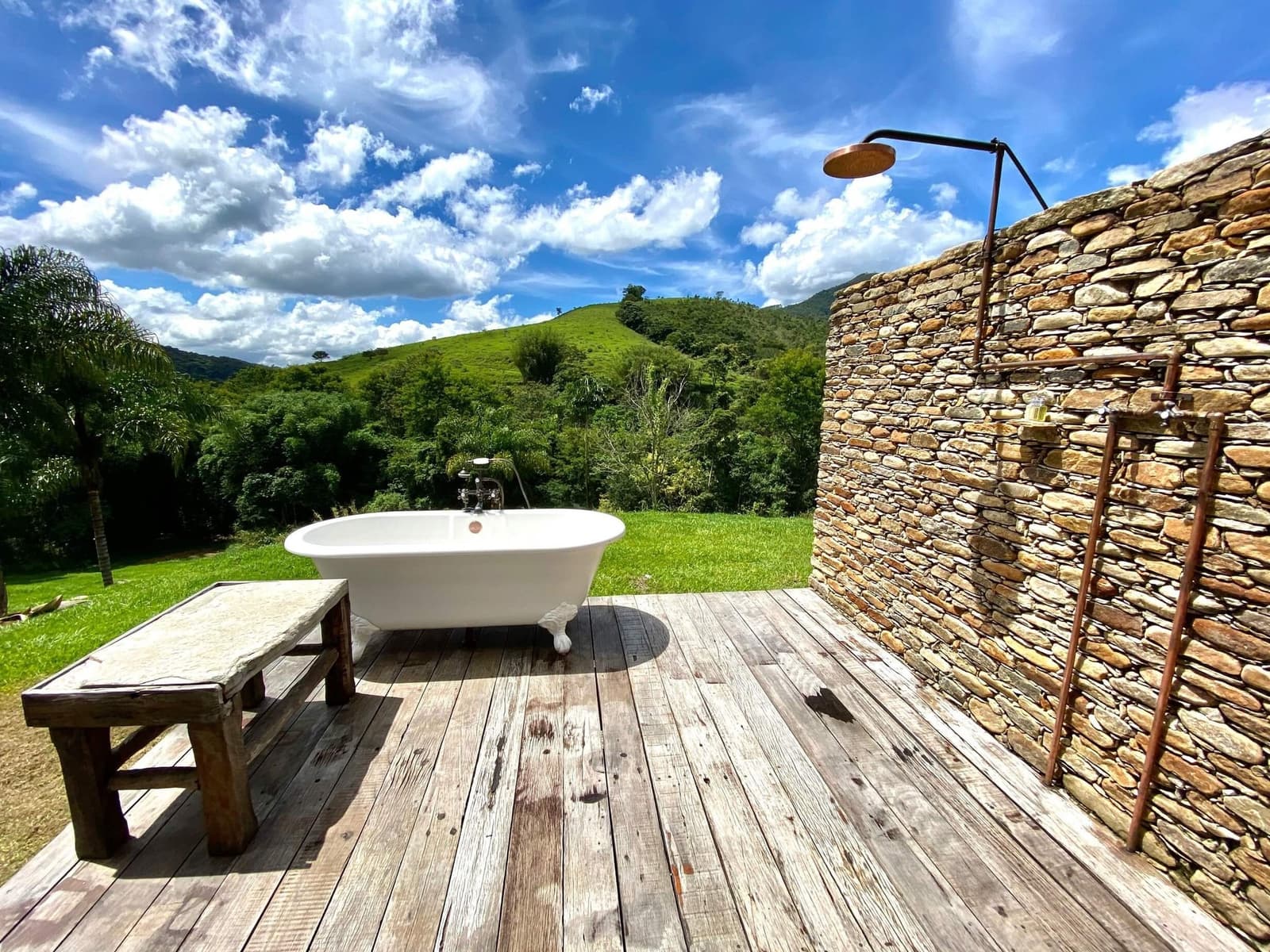 Your next bath comes with hills, clouds, and zero neighbours. A white clawfoot bathtub on a weathered timber deck at the Ibiti Project, with a copper outdoor shower beside a dry-stone wall and rolling green hills under a blue sky beyond